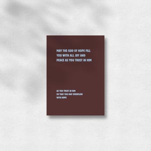 Könnte beinhalten: Ein rechteckiger, dunkelbrauner Druck mit hellblauem Text, der lautet: MAY THE GOD OF HOPE FILL YOU WITH ALL JOY AND PEACE AS YOU TRUST IN HIM AS YOU TRUST IN HIM SO THAT YOU MAY OVERFLOW WITH HOPE. Das Design ist mittig auf der Seite platziert.