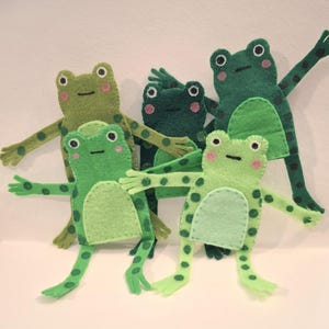May include: A group of six handmade felt frog figures in varying shades of green. Each frog has a light green belly, pink cheeks, and black dot details. The frogs have outstretched arms and legs, giving them a playful appearance.