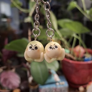 May include: A pair of handmade earrings with small, grumpy-faced, pale yellow figures. The figures have black eyes and are suspended from silver-coloured chain links. The background is a blurred view of green plants and a red pot.