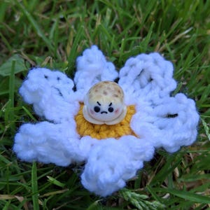 May include: A handmade crochet flower with white petals and a yellow center. The flower features a small, round, grumpy-faced character in the center. The item is set against a backdrop of green grass.