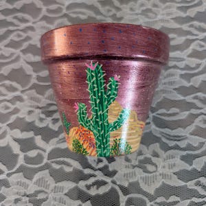 May include: A small, hand-painted terracotta flower pot with a metallic rose-gold finish. The pot features a vibrant desert scene with a green cactus, orange and yellow desert plants, and pink flowers. Blue dots are scattered across the rim.
