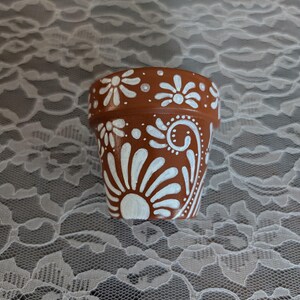 May include: A small, terracotta-colored flower pot decorated with white floral and swirl designs. The hand-painted pot features a sunburst pattern and other stylized flowers. The pot is likely used for planting small flowers or herbs.