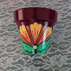 Puede incluir: Maceta de cerámica de color burdeos con un diseño floral pintado a mano. La flor presenta pétalos amarillos, detalles en naranja y rojo, y hojas verdes. Adecuada para uso en interiores o exteriores.