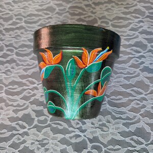 May include: A dark green painted flower pot with orange, blue, and green floral designs. The pot has a metallic sheen and a wide opening. The floral design features bird of paradise flowers.