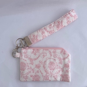 Op de afbeelding: Een bijpassende set van een kleine etui met ritssluiting en een polsbandje. Beide items zijn gemaakt van witte stof met een roze bloemenpatroon. De etui heeft een roze rits en een zilverkleurige metalen sluiting.