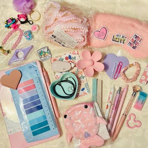 Peut inclure: Une collection de papeterie et d'accessoires comprenant un masque de nuit rose en peluche avec des autocollants, un plat en forme de cœur, un carnet floral, des stylos, une règle avec des onglets adhésifs, une pochette rose en forme de poisson.