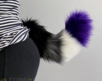 May include: A plush, faux fur animal tail featuring a gradient color pattern that transitions from black at the base to gray, then white, and finally a vibrant purple tip. The tail is attached to a black garment and has a thick, fluffy texture.