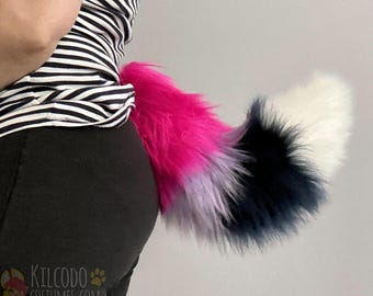 May include: A long, plush faux fur tail featuring a color-blocked design with sections of vibrant hot pink, light lavender, deep navy blue, and crisp white. The tail is attached to a black garment and displays the text Kilcodo Costumes.com.