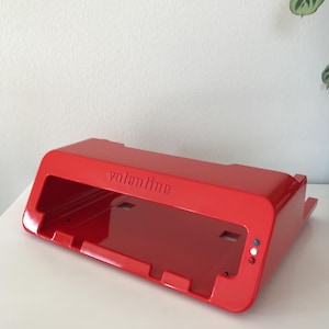 Peut inclure: Un objet rectangulaire rouge vif avec le mot "valentine" en relief sur le dessus. L'objet a un design incliné avec une zone encastrée et de petites lumières circulaires sur le côté. Il repose sur une surface blanche.