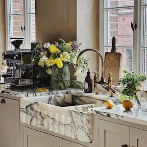 May include: A kitchen with a marble countertop and a farmhouse sink. A brass tap and a vase of yellow and purple flowers are on the counter. A black espresso machine is on the left, and a wooden chopping board leans against the window.