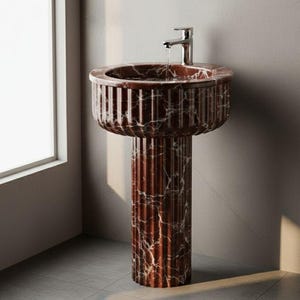 May include: A freestanding pedestal sink made of red marble with white veining. The sink has a round basin with a fluted rim and a tall, cylindrical pedestal. A chrome faucet is mounted on the sink, with water flowing into the basin.