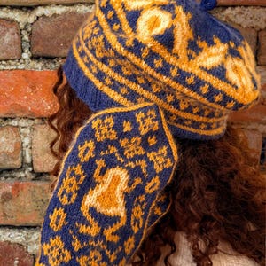 May include: A knitted blue and yellow beret and scarf set. The beret and scarf feature a detailed pattern in blue and yellow. The beret has a small pom-pom on top. The scarf is wrapped around the person's arm.