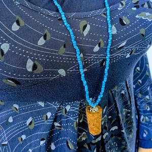 May include: A necklace with a turquoise beaded chain and a cork pendant. The pendant is a light brown color and has a decorative metal cap. The necklace is worn over a dark blue dress with a white and green pattern.