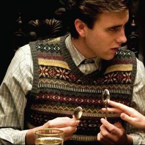 Puede incluir: Una persona con un chaleco gris estampado sobre una camisa con cuello. El chaleco presenta un diseño geométrico en tonos rojos, amarillos, verdes y blancos. La persona sostiene dos cucharas y un vaso.