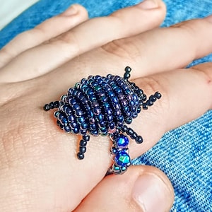 Peut inclure: Bague en forme de scarabée, faite de petites perles bleu foncé avec des reflets irisés. Le scarabée a des pattes et des antennes noires, et un groupe de perles bleues sur la bande de la bague. Bijou original.
