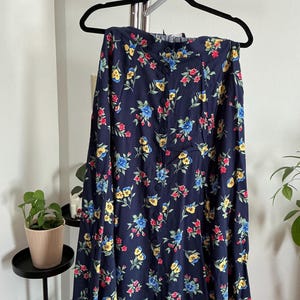 May include: A navy blue floral print skirt with a button-down front. The skirt features a pattern of red, yellow, and blue flowers. The skirt is hanging on a black hanger.