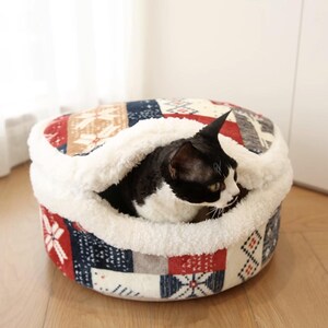 May include: A cozy cat bed with a patchwork design in red, navy, and cream colors. The bed has a soft, white, plush interior and a round shape. A black and white cat is nestled inside the bed.