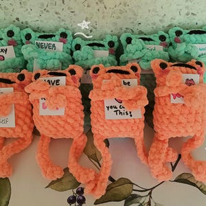 May include: A collection of handmade crochet frog plushies in shades of orange and teal. Each frog has a unique expression and holds a small card with motivational phrases like "Never" and "Believe in yourself."