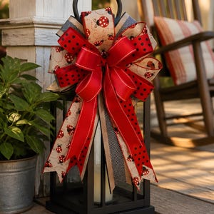 May include: Black metal lantern with a decorative bow. The bow features red, black polka dot, and ladybug patterned ribbons. A red bow is centered on the lantern. A candle is inside the lantern. The lantern is on a wooden deck.