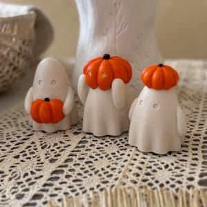 Könnte beinhalten: Drei weiße Geisterfiguren mit orangefarbenen Kürbisköpfen. Die Geister haben einfache, runde Augen und sind spielerisch dargestellt. Die Kürbisse sind leuchtend orange und verleihen eine festliche Note. Ideal für Halloween-Dekorationen.
