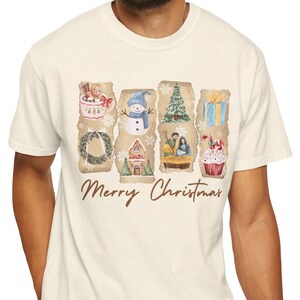 May include: Cream-colored t-shirt featuring a watercolor design of Christmas-themed images, including a wreath, snowman, and gifts. The words "Merry Christmas" are printed in a script font below the images. The shirt is made of a soft material.