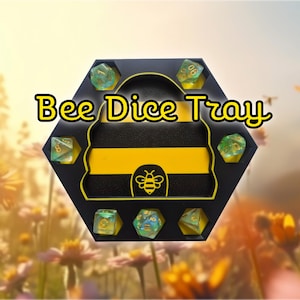 May include: A black and yellow hexagonal dice tray with the text "Bee Dice Tray". The tray has a bee design and holds seven green and gold dice. The background is a blurred image of flowers.