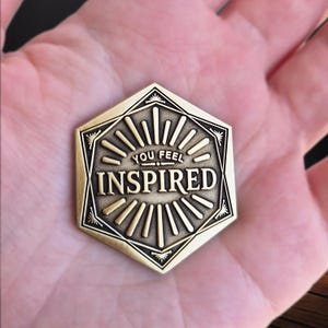 May include: A bronze-colored, hexagonal coin with the words "YOU FEEL INSPIRED" in a stylized font. The coin features a sunburst design and decorative borders. The coin is held in a hand.