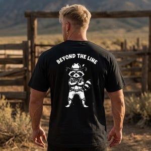 Puede incluir: Camiseta negra con un gráfico blanco de un mapache con sombrero de vaquero y el texto "BEYOND THE LINE". El mapache tiene los brazos cruzados y una cola a rayas. La camiseta está hecha de un material suave.