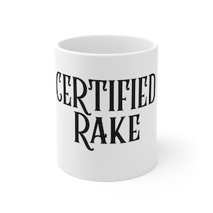 Könnte beinhalten: Weißer Keramikbecher mit dem schwarzen Schriftzug "CERTIFIED RAKE". Die Schrift ist in einem dekorativen Stil gehalten. Der Becher hat eine zylindrische Form.