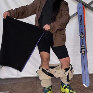 May include: A brown hooded jacket, black shirt, black shorts, and beige pants worn around the ankles with neon yellow ski boots. A black rectangular fabric piece is held up, and a purple Voile Vector 179 cm ski stands vertically nearby.