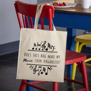 May include: A cream-colored tote bag with black musical notes and the text "DOES THIS BAG MAKE MY MUSIC LOOK ORGANIZED?" is hanging on a red chair. The background includes a kitchen with a blue table and a plate of strawberries.