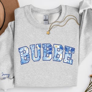 May include: A light gray sweatshirt with the word "BUBBE" in blue letters with a floral design. The sweatshirt has a crew neck and long sleeves. A gold necklace with a star pendant is draped across the sweatshirt.