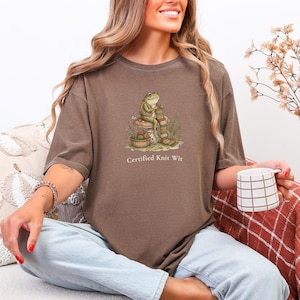 May include: A brown short-sleeve t-shirt featuring a graphic of a frog sitting on a mushroom, knitting a striped scarf with a basket of yarn nearby. The text below the graphic reads, "Certified Knit Wit" in a simple, light-colored font.