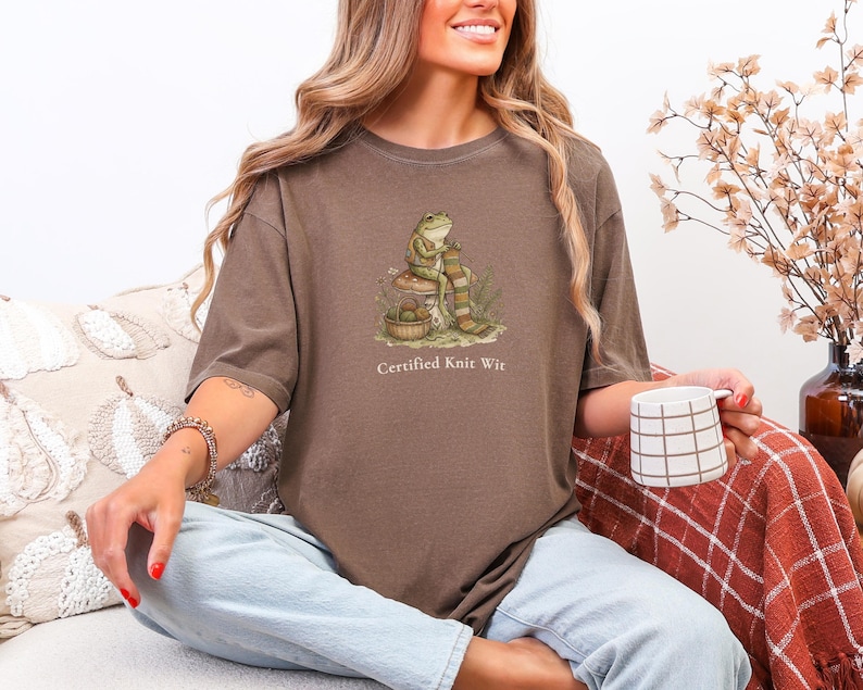May include: A brown short-sleeve t-shirt featuring a graphic of a frog sitting on a mushroom, knitting a striped scarf with a basket of yarn nearby. The text below the graphic reads, "Certified Knit Wit" in a simple, light-colored font.