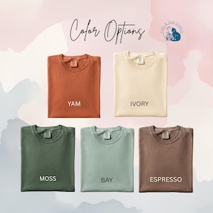 May include: Five folded t-shirts in various colors: yam, ivory, moss, bay, and espresso. The text "Color Options" is written in cursive at the top, with a logo in the upper right corner. The background has a watercolor effect.