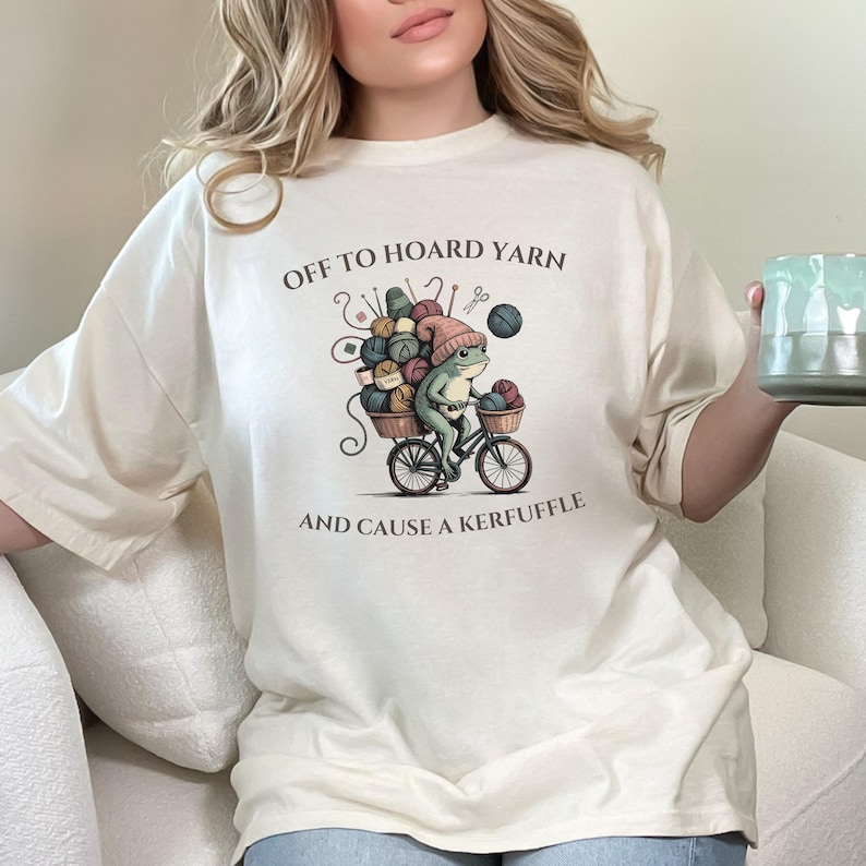 May include: Cream-colored t-shirt featuring a whimsical illustration of a frog riding a bicycle loaded with yarn. The text "OFF TO HOARD YARN AND CAUSE A KERFUFFLE" is printed above and below the graphic. The frog wears a pink hat.
