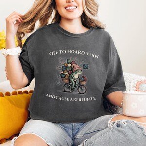 May include: Dark gray t-shirt with the text "OFF TO HOARD YARN AND CAUSE A KERFUFFLE". The shirt features a frog riding a bicycle loaded with yarn. The frog wears a pink hat and carries a basket of yarn.