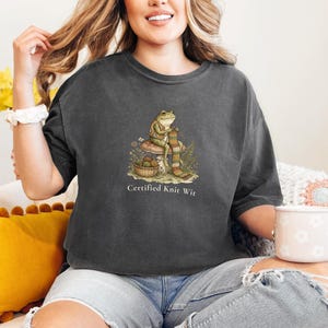 May include: Dark gray t-shirt featuring a whimsical illustration of a frog knitting a scarf, with the text "Certified Knit Wit" below. The frog is seated on a mushroom, with a basket of yarn nearby. The shirt is made of soft material.