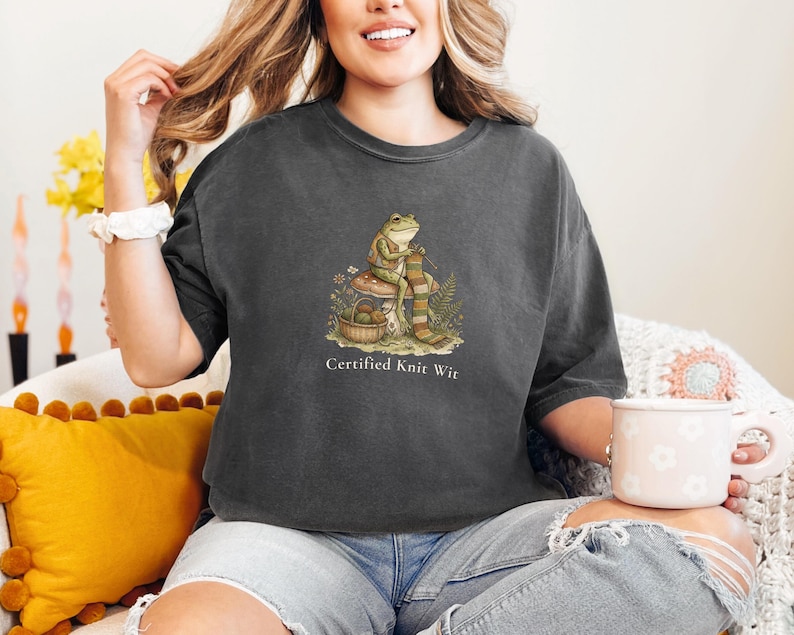 May include: Dark gray t-shirt featuring a whimsical illustration of a frog knitting a scarf, with the text "Certified Knit Wit" below. The frog is seated on a mushroom, with a basket of yarn nearby. The shirt is made of soft material.
