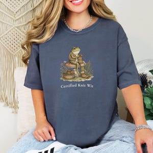 May include: A slate blue short-sleeve t-shirt featuring a graphic of a frog sitting on a mushroom, knitting a striped scarf with a basket of yarn nearby. Below the illustration, the text reads Certified Knit Wit in white lettering.