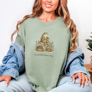 May include: Sage green t-shirt featuring a whimsical illustration of a frog knitting on a mushroom. The frog is surrounded by foliage and a basket of yarn. The text "Certified Knit Wit" is printed below the graphic.