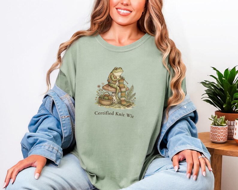May include: Sage green t-shirt featuring a whimsical illustration of a frog knitting on a mushroom. The frog is surrounded by foliage and a basket of yarn. The text "Certified Knit Wit" is printed below the graphic.