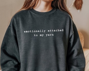 May include: Dark gray sweatshirt with the text "emotionally attached to my yarn" in white. The sweatshirt is a crew neck style and appears to be made of a soft material. The image shows the upper body of a person wearing the sweatshirt.