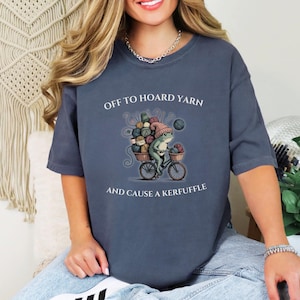 May include: A blue-grey t-shirt features a whimsical illustration of a frog riding a bicycle laden with yarn. The text reads "OFF TO HOARD YARN AND CAUSE A KERFUFFLE". The frog wears a pink hat and the bicycle has a basket of yarn.