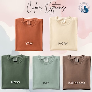 May include: Five folded t-shirts in various colors: yam, ivory, moss, bay, and espresso. The text "Color Options" is written in cursive at the top. The brand logo is in the upper right corner. The shirts are displayed on a light pink background.