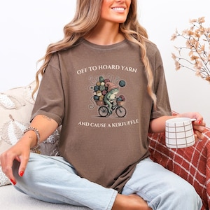 May include: Brown t-shirt with the text "OFF TO HOARD YARN AND CAUSE A KERFUFFLE". The graphic shows a frog riding a bicycle loaded with yarn. The frog wears a hat and is holding a ball of yarn. The shirt is a neutral color.