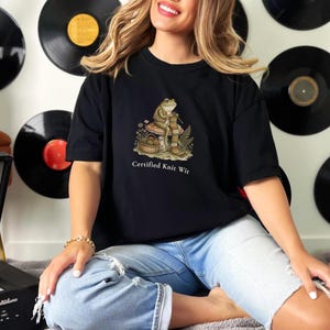 May include: Black t-shirt featuring a whimsical illustration of a frog knitting, seated on a mushroom, with the text "Certified Knit Wit." The design is in shades of brown, green, and beige. The shirt is a casual, short-sleeved style.