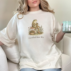 May include: Cream-colored t-shirt featuring a whimsical illustration of a frog knitting on a mushroom, with the text "Certified Knit Wit" below. The frog is knitting a striped scarf. A green and gray mug is held in the hand.