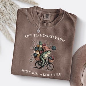 May include: Brown t-shirt with the text "OFF TO HOARD YARN AND CAUSE A KERFUFFLE". The graphic shows a frog riding a bicycle loaded with yarn. A silver necklace is draped on the shirt.