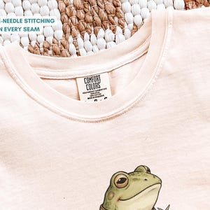 May include: A light peach-colored t-shirt with a crew neck and a frog graphic. The shirt has the text "DOUBLE-NEEDLE STITCHING ON EVERY SEAM" and a Comfort Colors tag. The frog is green with a brown collar.